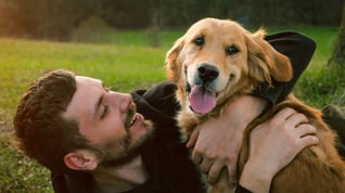 A man lying on grass smiling up at a happy golden dog who is leaning over him with its tongue out, as they play outdoors.